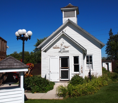 [White clapboard building with short tower at the top and a smaller section attached to front with doorway. Bell in a small structure on the grounds in front of the sidewalk near the school building.]